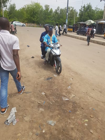 The image depicts a street scene in Dabawa, Nigeria. Two men ride a black and silver motorcycle. The driver wears a blue garment, while the passenger behind him wears a dark garment. A man in blue jeans and a white striped shirt stands in the foreground, looking down at his phone. The street and surrounding dirt area are littered with debris. In the background, there are other people, vehicles, trees, and a storefront.
