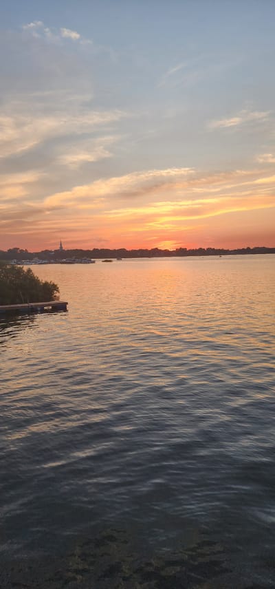 The image depicts a serene sunset over a body of water, likely a river or lake, with a distant tree line and a faint silhouette of a building with a steeple. The sky is a gradient of soft blues and oranges, with wispy clouds catching the last rays of the sun. In the foreground, the water exhibits gentle ripples, reflecting the warm hues of the sunset. To the left, a small wooden dock with dark blue floats is visible, partially obscured by lush green foliage. A few boats are scattered across the water in the mid-ground, near the shore. The overall impression is one of tranquility and natural beauty. This scene is evocative of a quiet evening on the Richelieu River in Chambly, Canada, known for its picturesque waterfront and historical landmarks like the Chambly Canal and Fort Chambly.