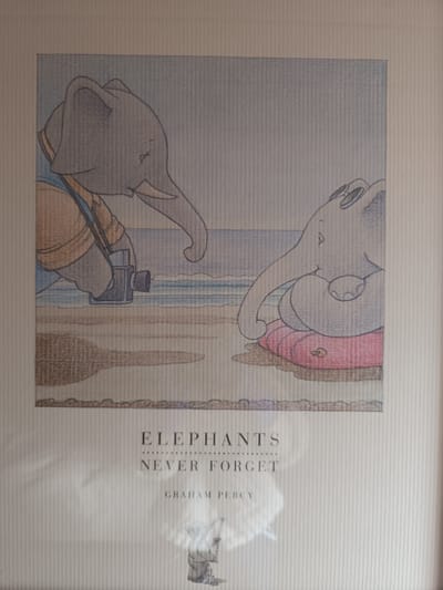 The image is a framed illustration featuring two cartoon elephants on a beach. One elephant, on the left, is wearing an orange vest and holding a camera, appearing to take a photo. The other elephant, on the right, is sitting on a pink inflatable ring, wearing sunglasses, and looking towards the left. The background depicts a beach scene with gentle waves and a pale sky, suggesting a calm, daytime setting. The overall tone is whimsical and serene.

Below the illustration, there is visible text. The largest text reads "ELEPHANTS" in bold, uppercase letters. Below this, a dotted line separates it from the text "NEVER FORGET", also in uppercase letters. Finally, the name "GRAHAM PERCY" is printed in smaller, uppercase letters. The illustration is done in a colored pencil or crayon style, with soft lines and muted colors. The textured paper of the print is visible through the framing. There are no specific cues to indicate the time of day or weather beyond the general beach setting.