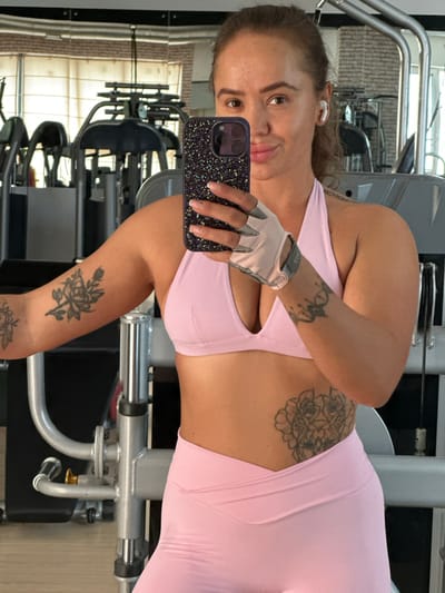 A young Caucasian woman with brown hair pulled back in a ponytail, fair skin, and brown eyes is taking a mirror selfie in a gym. She is wearing a pink sports bra and pink leggings. She has several tattoos: a rose bouquet on her upper left arm, a mandala-like design on her right forearm, and a floral design on her right hip. She is holding a black phone with a glittery case in her right hand and wearing a light-colored weightlifting glove on her left hand. She also has white wireless earbuds in her ears. The background shows gym equipment, including weight machines and mirrors. The lighting is bright and appears to be natural light coming from windows. The woman is looking directly at the camera with a slight smile. The image does not provide any information about the specific location in the city of Yuzhnouralsk, Russia.