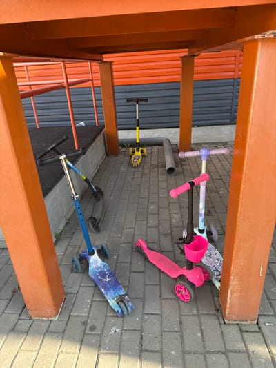 In Novoaltaysk, Russia, under an orange structure, several scooters are parked on a paved area. A blue and silver scooter with a cosmic design is in the foreground, leaning against a grey wall. To its right, a pink scooter with a basket is positioned next to a scooter with a purple handlebar and a white deck featuring a cat design. Further back, a yellow scooter with a black handlebar stands upright, and another dark scooter is partially visible behind the blue one. The environment appears to be an outdoor public space, possibly a playground or a recreational area, with a grey and orange striped wall in the background.