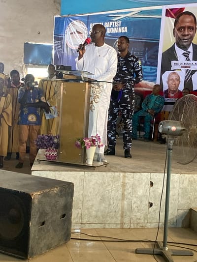 This image captures an indoor event taking place on an elevated stage within a structure in Ube, Nigeria, identified by a banner as 'Bethel Baptist Church Akwanga.' A dark-skinned male speaker, wearing glasses and a white long-sleeved traditional garment, stands centrally behind a clear acrylic and golden-based podium, holding a red-tipped microphone in his right hand. An open book is visible on the podium. To his right, a dark-skinned male in a blue/grey camouflage uniform stands upright, observing. On the stage's left, multiple individuals in light brown robes are present, one of whom, wearing a blue patterned shirt, holds a mobile phone, pointed towards the speaker. Behind the stage are large banners, one blue with a church emblem, and another displaying portraits, including 'Rev. Dr. Bulus A. Akwanga.' Two floral arrangements are positioned on the stage floor in front of the podium. Off-stage, a large black audio speaker box is in the lower left, and a grey standing fan with visible electrical cords is in the lower right. The stage features a tiled facade, and the background walls are light-colored and textured, with a bright light source visible on the far left.