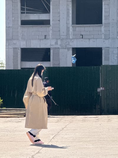 The image displays an outdoor scene, notably rotated 90 degrees counter-clockwise, showing a person standing near a construction site.

**Subjects:**
A person with long, dark hair is the central subject, positioned towards the right side of the rotated image. They are wearing a long, light-colored (beige or tan) coat and light-colored pants with a subtle pattern. On their feet are distinctive pink sneakers with black straps or laces. The person holds a smartphone in their raised left hand, seemingly using it for photography or videography. Behind them, a black pole, possibly part of a monopod or tripod, is visible.
The background features an unfinished multi-story building constructed from light grey concrete blocks or bricks, characterized by several dark, empty window openings. Within one opening, a blue object, possibly a tarp or bag, is discernible. Another opening shows internal structural elements.
Separating the person from the unfinished building is a tall, dark green barrier or wall, which appears to be a construction fence or tarp. A small, light-colored object with a red marking is attached to this barrier.
The ground where the person stands is a light-colored, textured concrete surface.

**Activity or event taking place:**
The person appears to be engaged in taking photos or videos with their smartphone, likely documenting the environment or themselves within the construction setting.

**Notable details:**
The scene is brightly lit by the sun, casting strong, defined shadows of the person and other objects onto the concrete ground. This indicates a clear, sunny day. The unfinished building and construction barrier are clear location cues, suggesting an active development or construction zone. The specified location context is Zhongtun, China. There is no visible text in the image.