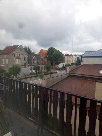 The image shows a view from a balcony looking out onto a street in Czersk, Poland. The sky is overcast with heavy clouds. In the foreground, a dark wooden railing with decorative cutouts obscures the lower part of the scene. Beyond the railing, across a paved street, are several buildings. On the left, two older, two-story houses with red tile roofs are visible. One is gray and has a bay window, while the other is a brighter red brick. Several trees are planted along the sidewalk in front of these houses. Further down the street, more modern buildings with light-colored facades and darker roofs are visible, including a tall radio tower in the distance. A black car is parked on the street. The environment appears to be a residential or mixed-use area within a town or city.