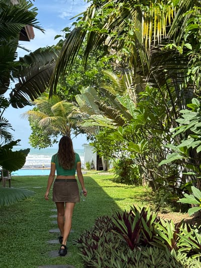 In Goviyapana, Sri Lanka, a woman with long brown hair, viewed from the back, walks on a stone path through a lush garden towards the ocean. She is wearing a green crop top with yellow trim and a brown A-line skirt. She carries a pair of glasses in her right hand. The garden features manicured grass, palm trees, and various tropical plants. In the background, a bright blue swimming pool is visible, and beyond that, the ocean meets the horizon under a clear sky with scattered clouds. The scene suggests a serene tropical resort setting.