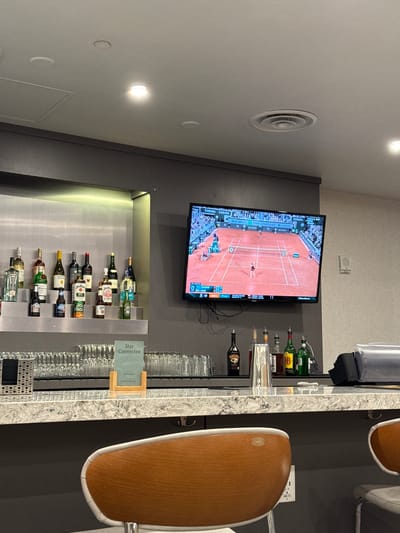 The image depicts a modern bar area, likely within an airport lounge. A large flat-screen television is mounted on a dark grey wall, displaying a tennis match on a clay court. The screen shows "BNP PARIBAS" sponsorship, "FINAL" status, player names including "SABALENKA," and game scores. "Emirates" branding is also visible on the TV screen.
Below the television, various liquor bottles are arranged on illuminated shelves, including "JIM BEAM," "BAILEY'S," and a partially visible "COORS LIGHT." A light-colored granite bar counter runs along the right side of the frame, laden with rows of clean glassware. Two modern bar stools with light wood backs and white upholstered seats are visible, pulled up to the counter.
A small green sign on the bar countertop reads "Stay Connected" and includes "The Club MCO" and "Emirates," indicating the location is likely an airport lounge at Orlando International Airport (MCO). An electrical outlet on the wall behind the stools is labeled "LMC-21." Overhead lighting fixtures and a circular air vent are visible on the light grey ceiling, contributing to the indoor, contemporary setting. The scene suggests a leisure area for travelers to relax and watch sports.