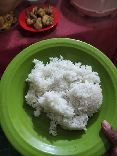 A green plate filled with white rice sits on a red tablecloth. The rice is steaming and appears to have been recently cooked. There is a partial view of a hand to the right of the plate and a blurred background showing two additional plates with food. The coordinates given are for a location in the city of Kecamatan Lawang, Indonesia. The image is likely taken indoors at a kitchen table.