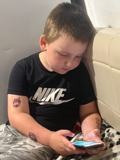 In an indoor setting, likely a bedroom, a young Caucasian boy with short brown hair is leaning against a light-colored headboard, looking down at a smartphone he is holding in his hands. He is wearing a black Nike t-shirt and camouflage patterned pants. The boy has several temporary tattoos on his right arm, including a wolf's head on his upper arm and what appears to be a cluster of circles on his forearm. The image is a medium shot, focusing on the boy and his immediate surroundings. There is no indication of the boy's location within the city of Chelyabinsk, Russia.
