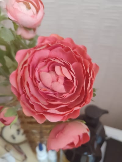 A close-up, slightly blurred image shows a bouquet of artificial pink ranunculus flowers. The main focus is on a large, fully bloomed flower in the center, with its layered petals displaying a gradient of pink hues. To the left and bottom, there are other partially visible buds and flowers, also in shades of pink. Green leaves are scattered amongst the blooms. In the background, a woven basket can be seen, and to the right, a dark, possibly ceramic, dispenser with a curved spout is partially visible, along with a blurred white bottle. The overall lighting is soft, and the background is a neutral, textured gray wall. The image does not provide any contextual information about its location within the city of Donetsk, Ukraine.