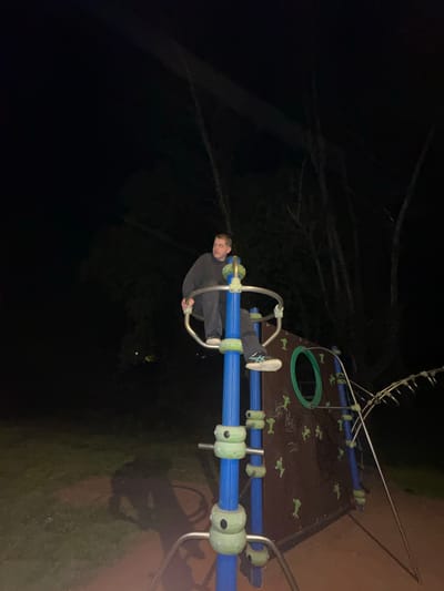 A nighttime photograph depicts a single male subject positioned on playground equipment within a park in Guimarães, Portugal. The male, appearing to be a young adult, is seated atop a blue and light-green tubular structure, gripping silver bars with both hands. He is dressed in a dark grey long-sleeved shirt, dark trousers, and sneakers with white soles. Behind him, a brown climbing wall is visible, featuring multiple green handholds, some shaped like figures, and a prominent circular green opening. To the subject's right, a metallic, ladder-like structure extends diagonally upwards. The ground beneath the equipment comprises a reddish-brown soft surface and patches of dark grass, with shadows cast by the playground elements. The background is characterized by silhouetted trees with visible branches against a dark, unlit sky.