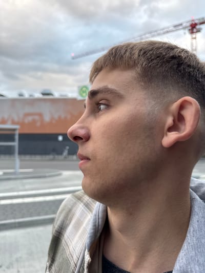 The image shows a close-up, profile view of a young Caucasian male with light brown hair and a light complexion, looking to the left. He is wearing a plaid collared shirt over a gray hooded sweatshirt. In the background, there is a blurred urban environment with a large orange building, a gray structure, and a construction crane with a red top. The sky is overcast with clouds. The overall setting suggests an outdoor location in a developed area, possibly a city. Based on the context of Lörrach, Germany, this scene could be depicted near the construction site of a new building or infrastructure project within the city.
