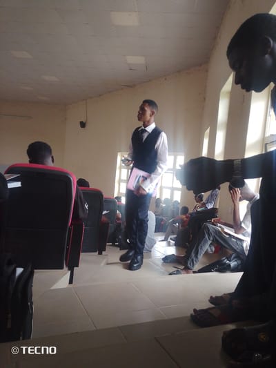 This image depicts a group of young men in what appears to be an indoor educational setting, possibly a classroom or lecture hall, located in Gumel, Nigeria. The room has light-colored walls and a tiled floor. Sunlight streams in from windows, creating distinct shadows.
In the foreground, a young man in a white shirt and dark vest stands facing left, holding a pink folder. To his left, another young man sits with his back to the camera, wearing a black shirt. Behind him, rows of dark chairs with red accents are visible.
Further back, several other young men are seated, some appearing to be engaged in a lesson or discussion. One individual in the middle ground is seen wearing a white shirt and dark pants, with his arm raised and hand near his head, as if in thought or speaking. On the far right, a person's legs are visible, wearing dark pants and black shoes. The overall atmosphere suggests a formal or academic gathering. The presence of the "TECNO" logo in the bottom left corner indicates that the image was taken with a Tecno mobile device.