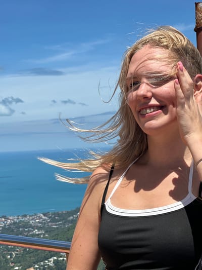 A blonde woman with braces, wearing a black tank top with white trim, smiles at the camera on a windy day. Her hair is blowing across her face, and she has her hand near her temple. She is standing on a balcony or railing overlooking a city and the ocean. The city is likely Ko Samui, Thailand, given the tropical vegetation and architecture visible in the background. The sky is blue with scattered clouds.