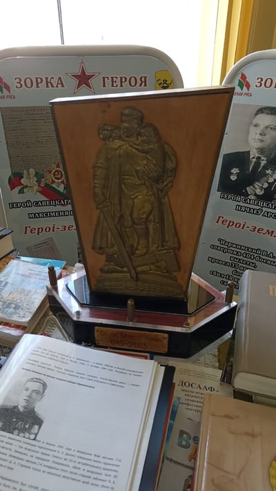 The image depicts a display in Kapyĺ, Belarus, featuring a wooden trophy with a relief of a soldier embracing a child. The trophy is positioned on a pedestal with a plaque reading "60 лет Великой Победы 1945-2005". Surrounding the trophy are several books and banners. One banner on the left is partially visible and contains text in Belarusian, with the word "ЗОРКА" (star) prominently displayed. Another banner on the right also shows Belarusian text and a portrait of a decorated soldier. In the foreground, an open book displays a black and white photograph of a young man in military uniform, accompanied by text in Belarusian detailing his military service. The environment appears to be an indoor setting, possibly a museum or a commemorative exhibition, with a bright light source suggesting a window or overhead lighting. The overall scene commemorates the 60th anniversary of Victory Day and highlights heroes of the Great Patriotic War.