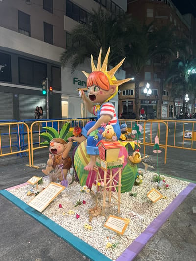 The image depicts a street scene at night in Alicante, Spain, likely during a festival given the elaborate display. In the foreground, a large, brightly colored sculpture of a cartoonish child with a sun-like headdress is the main subject. The child is seated on a giant, striped egg-shaped object, holding a magnifying glass up to their eye. To the left of the child is a smaller, brown, furry creature with large eyes and an open mouth, sitting next to a purple shovel. This creature appears to be reaching for the child's hand.
The sculpture is situated on a bed of white pebbles, interspersed with small artificial flowers and foliage. There are several small, yellow signs with text in Spanish placed around the base of the sculpture, some of which are tilted. Behind the sculpture, yellow and blue barricades cordon off the display from the street.
In the background, palm trees are visible, along with the facades of buildings. One building has a prominent sign for "Farma" which is partially obscured, and another sign for "Colchones." A traffic light with a green light illuminated is visible on the left side of the image, and streetlights illuminate the scene. Two individuals are standing near the traffic light, observing the display. The overall impression is of a festive, nighttime event in a public urban space.