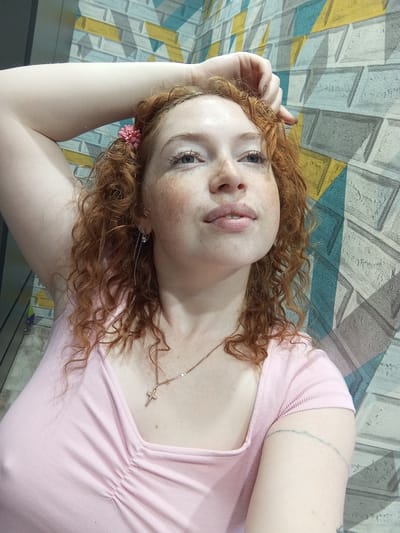 A young woman with red curly hair lies on a surface with a geometric pattern of gray, blue, and yellow. She is wearing a light pink top and a delicate gold necklace with a cross pendant. A small pink flower is tucked into her hair near her ear. She has fair skin with freckles and is looking towards the camera. A faint circular tattoo is visible on her left arm. The image appears to be taken indoors.
The location in Yurgamysh, Russia, cannot be determined from the image.