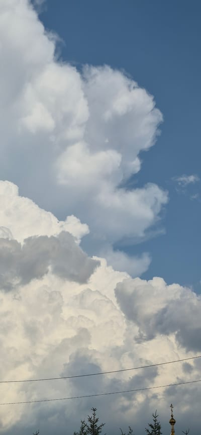 The media file is a low-angle shot of a dramatic sky filled with large, billowing cumulus clouds against a bright blue sky. The clouds are illuminated by sunlight, with some areas appearing bright white and others showing shades of gray, suggesting depth and volume. The composition is dominated by the clouds, which fill most of the frame, creating a sense of awe and grandeur.
In the upper right portion of the image, there are thin, dark wires running vertically, characteristic of power or utility lines. Also in the upper right, a portion of a decorative architectural element, possibly a spire or finial with intricate detailing and some gold coloring, is visible, along with dark green foliage from a tree. These elements suggest a human-made landscape intersecting with the natural sky.
The overall impression is of a bright, possibly late morning or afternoon sky, with dynamic cloud formations. The scene is serene yet powerful, with the interplay of light and shadow on the clouds adding to the visual drama. The presence of utility lines and architectural detail grounds the sky in a specific, potentially urban or semi-urban, environment. The location context of Ussurijsk, Russia, adds a geographical marker to the scene. There is no visible text within the image.