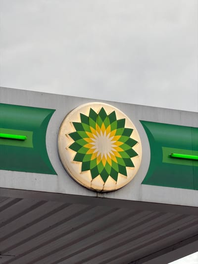 An outdoor close-up depicts the upper section of a BP gas station canopy in Vila Real, Portugal, viewed against an overcast, light grey sky. The central feature is a large, circular BP 'helios' logo, showcasing its characteristic green, yellow, and white petal design. The logo is internally lit, casting a golden glow around its perimeter, and is mounted on a light grey horizontal fascia panel. This fascia incorporates dark green accent panels with curved lighter green elements. Two prominent, horizontally aligned bright green neon or LED tube lights are integrated into these green sections, positioned to the left and right of the main BP emblem. The ribbed, grey underside of the canopy is visible in the lower portion of the frame. The entire structure exhibits visible signs of weathering and dirt accumulation. No individuals, vehicles, or active elements are present in the scene.