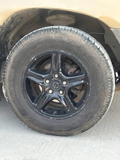 The image displays a close-up of a dirty, black car wheel and tire, likely belonging to a sport utility vehicle or van given the tire specifications. The vehicle's light-colored (possibly beige or cream) fender and a dusty wheel well are visible on the left. The setting appears to be outdoors, on a light grey, possibly concrete or asphalt, surface.
The wheel is a 5-spoke design, painted black, but shows considerable chipping and wear, revealing a silver color underneath. A silver Lexus logo is prominently featured on the center cap. Behind the wheel spokes, disc brakes (rotor and caliper) are visible, appearing dirty and somewhat corroded.
The tire is clearly labeled as an M+S (Mud + Snow) type, indicating its suitability for various weather conditions. Its size and specifications are "235/65R16C 115/113R BAR". Brand names "VAN TRAX" and "JOYROAD" are visible on the sidewall. Additional text includes "TUBELESS", "STEEL BELTED RADIAL", and details about its ply construction: "TREAD POLYESTER 2 STEEL 2+ NYLON" and "SIDEWALL POLYESTER 2". A partial "SAFETY WARNING: SERIOUS" is also present. The tire's tread shows signs of use and is covered in dust and some white residue, suggesting the vehicle operates in dusty conditions. No people are present, and no specific activity is taking place; it's a static shot of a car component in Epe, Nigeria.