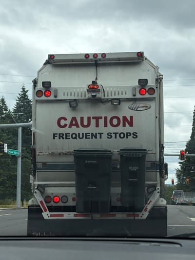 The image shows the rear of a white garbage truck with red lettering reading "CAUTION FREQUENT STOPS" on its back. The truck is equipped with two dark green garbage bins and multiple red brake lights. The truck is on a street with trees and a traffic light visible in the background. A street sign reads "SW Cedar Hills Blvd". The scene appears to be in Beaverton, United States of America. The sky is cloudy.