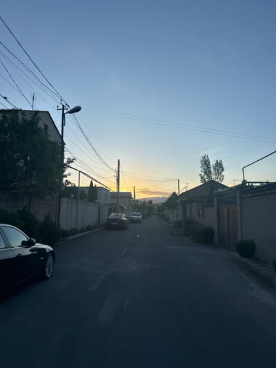The image depicts a street scene in Arinj, Armenia. The street runs horizontally, with buildings lining both sides. Several cars are parked along the street, including a black car in the foreground. Overhead power lines crisscross the sky. The sky is light blue near the horizon and deepens to a darker blue towards the top of the frame. The sun is setting in the distance, casting a golden glow on the horizon.