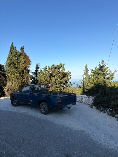 A blue pickup truck is parked on the side of a road in the Municipal Unit of Lefkada, Greece. The road is made of gray asphalt and is bordered by green trees and bushes. The sky is clear blue. The truck has a license plate that reads "EXA 7184." The truck appears to be old and in poor condition. The sea is visible in the background.