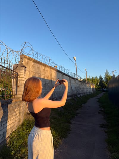 The image captures an outdoor scene in Новинки, Russia, during the golden hour, indicated by warm, low-angle sunlight and elongated shadows. A young female, seen from behind and slightly to the right, stands on the left side of a narrow, dark paved path. She has auburn bobbed hair and wears a black strapless top and light grey sweatpants with a visible pocket on the right leg. She is actively holding a dark-colored smartphone horizontally with both hands, seemingly photographing an area towards the right mid-ground.
To the subject's left, a light-colored brick pillar transitions into a dark metal fence with vertical bars, behind which green foliage is visible. Dominating the right side of the frame is a long, tall, weathered concrete wall, extending into the distance and topped with multiple coils of barbed wire. The paved path runs parallel to this wall, bordered by unkempt grass and weeds on both sides. Further along the path, two utility poles with luminaires (streetlights) are visible, and dense green trees rise above the concrete wall in the mid-ground. A single dark electrical cable crosses the clear, bright blue sky diagonally in the upper left portion of the image. Shadows from the wall and the subject extend across the ground, contrasting with the sunlit sections.