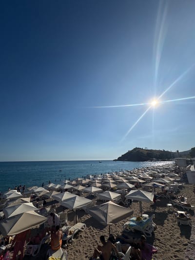 The image captures a crowded beach in Sutomore, Montenegro, under a clear blue sky with the sun shining brightly. The foreground is dominated by a sandy beach with numerous white and beige beach umbrellas set up, casting shadows on the sand. Beach chairs are arranged under and around the umbrellas, with many people lounging on them. Several individuals are visible on the beach, some sitting, some standing, and a few are partially obscured by umbrellas.

The middle ground features the turquoise sea, dotted with people swimming and enjoying the water. A few small boats are visible on the water, and in the distance, a small peninsula or island rises from the sea, covered in green vegetation and topped with some structures. The sun is positioned high in the sky, to the right of the peninsula, and its rays are visible as lens flares extending across the frame. The overall impression is of a busy, sunny day at a popular seaside resort.
