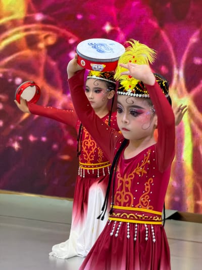 Two young girls in traditional red and white costumes are performing a dance on a stage in front of a vibrant pink and yellow background. They are wearing elaborate headdresses, with one girl sporting a bright yellow feather plume. Both girls are holding small tambourines. The girl in the foreground, positioned to the right, has her right arm raised and is looking forward, while the girl behind her, to the left, is also holding her tambourine up. The costumes feature ornate golden patterns and beaded embellishments. The background appears to be a large screen displaying a colorful, abstract design with glowing effects. The stage floor is a muted gray. The overall scene suggests a cultural performance or festival, possibly within the Jiulongpo District of China.