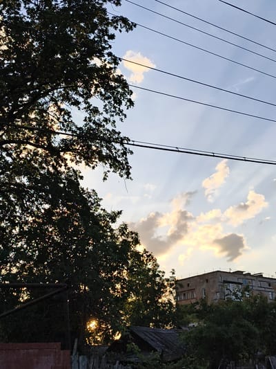 This image depicts an outdoor scene in Donetsk, Ukraine, during what appears to be late afternoon or early evening, as indicated by the soft, warm light and the crepuscular rays emanating from behind clouds. The sky is a pale blue, streaked with wispy clouds that are tinged with yellow and orange from the setting sun. Power lines stretch across the upper portion of the frame.
In the foreground, the dark silhouette of a large tree dominates the left side of the image, its dense foliage partially obscuring the sky and the light behind it. The setting sun is visible as a bright, shimmering orb peeking through the leaves. Beneath the tree, a section of a rusty metal fence and what looks like a dilapidated wooden fence can be seen, suggesting a somewhat rural or aged urban environment.
To the right, a nondescript, multi-story building with weathered concrete walls stands partially obscured by the surrounding greenery. The building has numerous windows, some appearing boarded up or covered with graffiti. The overall impression is one of a quiet, possibly slightly neglected, suburban or urban fringe area as the day draws to a close. There are no people visible in the image. The mood is somewhat serene, with a hint of melancholy due to the aging structures and the fading light.