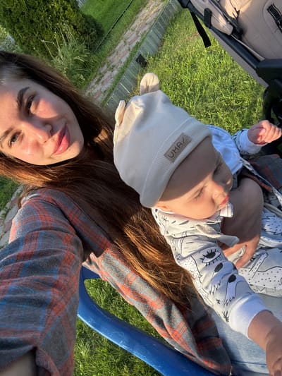 The image shows a woman and a baby outdoors, with grass and what appears to be a park or backyard in the background. The scene is bathed in the warm light of what seems to be late afternoon or early evening. The woman is in the foreground, looking towards the camera with a gentle smile. She has long, brown hair and is wearing a plaid shirt. The baby is lying on the woman's chest or lap, facing away from the camera and looking towards the left. The baby is wearing a white beanie with a small label that appears to have some text on it, possibly "UM&?" and a cartoonish design on its onesie. The overall mood of the image is peaceful and affectionate, suggesting a moment of quiet bonding between mother and child. The lush green grass and the golden sunlight indicate a pleasant, likely warm, day. The text "UM&?" is visible on the baby's beanie.