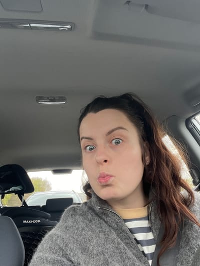 A young woman is shown in a close-up selfie taken inside a vehicle. She has fair skin, dark hair with brown highlights, and light-colored eyes. Her lips are pursed slightly. She is wearing a grey sweater with a grey and white striped garment underneath. Part of a "Maxi-Cosi" child safety seat is visible in the background. The interior of the car, including the ceiling and dome light, is visible. Based on the presence of the Maxi-Cosi child seat, it is likely she is either a parent or a caregiver. The location, Meisenheim, Germany, cannot be verified from the image.