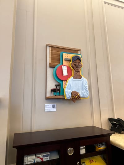 The image shows an art installation titled "Contemplation Chair" by Dada Khanyisa (2020) on a beige-painted wall. The artwork depicts a sculpted figure sitting at a desk. To the right of the artwork is a partially open brown wooden cabinet, showing a box of Cohiba cigars, a box of Stuyvesant cigarettes, and a cash register. The location appears to be in a building with architectural paneling, likely in Cape Town, South Africa, given the artist's identity.