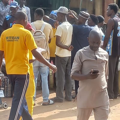 A medium shot depicts a group of approximately 12 men of varying heights and ages standing outside in Dadi Riba, Nigeria. The men are clustered closely together, with those in the foreground partially obscuring those behind. The man closest to the viewer is wearing a yellow polo shirt with the words "RTEAN KADUNA STATE" printed on the back, paired with dark blue track pants featuring white stripes down the sides. He is standing in front of a tire and an object with the text "ENG". The man to his left is wearing a light yellow shirt and has a tan satchel bag across his chest. Another man is wearing a grey shirt and pants. The background shows parts of buildings and structures, creating an urban setting. The ground appears to be dirt or a sandy color. One of the men in the group is holding a phone and appears to be taking a picture.