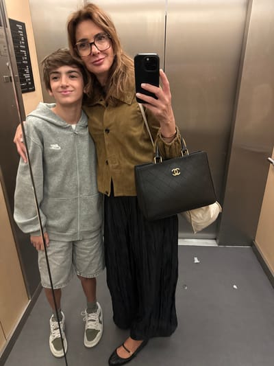 A woman and a child are in an elevator in Porto, Portugal. The woman, wearing glasses and a tan jacket, holds a smartphone to take a selfie. She carries a black Chanel purse. The child, wearing a gray Nike sweatsuit and sneakers, stands in front of her. The elevator interior is stainless steel with some signage visible. The floor is gray.