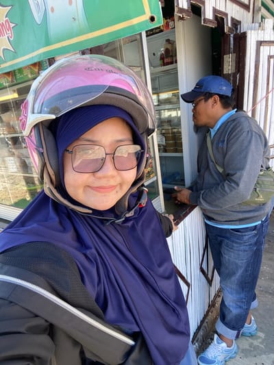 A woman wearing a purple hijab and a pink and white helmet, with square-framed glasses, is taking a selfie in the foreground. She is looking directly at the camera with a slight smile. Behind her, a man wearing a blue baseball cap, glasses, and a grey hooded sweatshirt over a blue shirt, is standing and interacting with a person off-camera. He has a green crossbody bag slung over his shoulder and is wearing blue jeans and light blue sneakers. They appear to be at an outdoor stall or shop with a refrigerator visible to the left, containing bottles and containers. The stall has a white wooden facade. The background suggests an outdoor setting, possibly a street in Sungai Paring, Indonesia, indicated by a green banner with text in Indonesian and a yellow border visible at the top of the image.