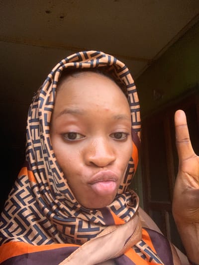 A selfie is captured from a medium close-up angle, with the subject, a young woman, in the foreground. She is wearing a patterned headscarf with a repeating geometric design in black and brown on an orange background. Her face is turned slightly towards the camera, with her lips pursed and her right hand raised in a peace sign. The background is dimly lit and appears to be indoors, with a dark, textured ceiling and a hint of green on the right side, suggesting a wall or an opening to the outside. The overall impression is of a candid moment captured in a simple, unadorned setting, possibly in Shagamu, Nigeria, given the context.