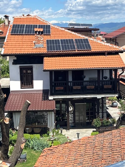 The image shows a building with a tiled roof, located in Bansko, Bulgaria. A section of the roof is fitted with solar panels. The building has a white facade with a window. The building also features a balcony with dark wood railings. Adjacent to the building is an outdoor area with plants and what looks like a patio, and a tree in the foreground with several branches cut. The sky is visible in the background, with clouds.