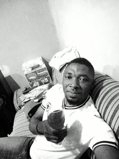 A black and white image displays a male individual, appearing to be an adult of African descent, seated indoors in Gwagwarwa, Nigeria. The man, positioned with a slight smile and direct gaze towards the camera, has short dark hair and a beard. He wears a light-colored polo shirt with dark stripes on the collar and sleeve cuffs, and dark trousers or jeans. His left hand holds a stemmed glass containing a dark liquid. He is leaning against a striped sofa or cushion. In the background, to his left, is a corner formed by two plain, light-colored walls. A carton, likely a product box with visible branding resembling "Minimie," is situated on a surface behind him, alongside various folded fabric items and a rolled textile object.