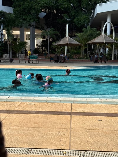 This image depicts an outdoor swimming pool area within a facility in Itirin, Nigeria, under natural daylight. In the immediate foreground, a light-colored, textured concrete pool deck with a drainage grate is visible. The midground features a blue swimming pool containing at least seven individuals, primarily children, partially submerged in the water. One child is visible in a pink top and a striped pink and white swim cap, while another wears goggles. Dark lane markings are discernible on the pool floor.

Adjacent to the pool, a person in a bright yellow shirt and dark pants stands near a multi-tiered trolley, possibly containing towels. Several empty wooden lounge chairs are arranged on the left side of the pool area. Further back, two distinct hexagonal umbrellas with slatted tops and white arched supports shelter multiple tables and chairs, under which groups of individuals are seated. The background consists of dense green foliage, including palm trees, and a multi-story building with prominent white architectural elements, columns, and balconies, visible through and above the trees.