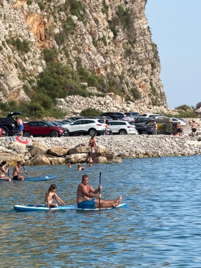The image depicts a coastal scene in Mirište, Montenegro, with a rocky cliff face rising in the background. A parking area with several cars is visible along the shore, situated on a rocky embankment. In the foreground, the clear blue water is populated with several people engaged in water activities. Two individuals are on a paddleboard in the middle ground, one adult male with a paddle, and a child seated in front of him. To the left, another paddleboarder is visible, and further to the left, two women are on a paddleboard. The water is calm with visible ripples. The overall impression is a sunny day at a popular beach or cove.