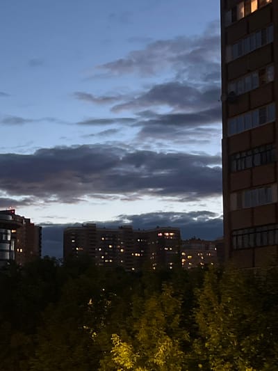 The image depicts a nighttime scene in Pushkino, Russia, with an urban landscape featuring buildings and trees. The orientation of the photo is vertical, showing a view from a lower vantage point looking upwards. In the foreground on the right, a large tree with dense foliage is visible, its leaves illuminated by artificial light, giving them a yellowish-green hue.

Behind the tree, several apartment buildings are seen. The windows of these buildings are lit, suggesting that it is evening or night. The architecture of the buildings appears to be typical of Soviet-era construction, with a repetitive facade. On the left side of the image, another building with a more modern, darker facade is partially visible, also showing illuminated windows.

The sky above is filled with dramatic clouds, which are dark and textured, suggesting a cloudy or stormy atmosphere. The contrast between the illuminated windows of the buildings and the dark sky creates a sense of depth and atmosphere. The overall mood of the image is somewhat somber, with the darkness and the textured clouds contributing to this feeling, yet the lit windows offer a touch of warmth and human presence within the urban environment. No text is visible in the image. The time of day is clearly night, and the weather appears to be cloudy.