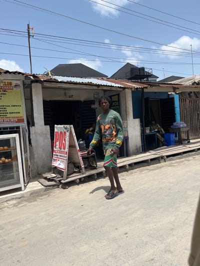 A person stands on a sidewalk in front of a building in Iranla, Nigeria. The person is wearing a green shirt with a print design and green and red shorts, and sandals. A sign that reads "POS SERVICES" is placed near the individual in front of a building that also has another sign reading "ELIS CAKES", indicating local businesses. The sidewalk is cracked and aged. Overhead, electrical wires are visible against a partially cloudy sky. In the background, another individual sits indoors on a chair.