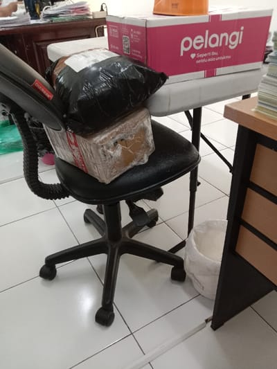 The image shows an office chair with a black seat and black wheels.  The chair is facing right and has a black armrest with the word "Chairman" printed on it. On the seat is a large cardboard box wrapped in clear plastic and a black bag that appears to be a packing bag. The chair is positioned under a white table with a brown wooden surface top. On the table is a pink box with the words "pelangi" and "Seperti Ibu, selalu ada untukmu" printed on it. There is a white fabric bucket beside the chair.  The floor appears to be white ceramic tile.  The location of the image is an office in Kecamatan Ngaliyan, Indonesia, based on the coordinates provided.