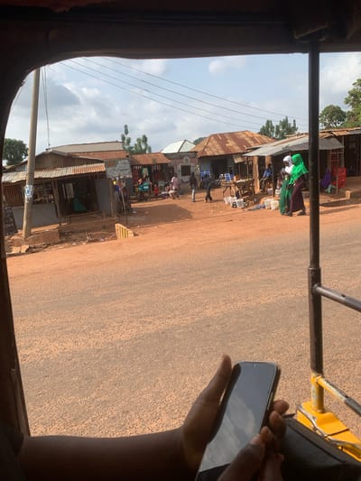 The image captures a street scene in Lafia, Nigeria, as viewed from inside a vehicle, likely an auto-rickshaw. The perspective is tilted, suggesting movement. The foreground is dominated by the interior of the vehicle, with a brown frame on the left and a portion of a yellow and black structure on the right, possibly part of the vehicle's framework. A hand holding a smartphone is visible on the right side, with the screen reflecting a cloudy sky.
The scene outside the vehicle features a dusty, unpaved road running parallel to a row of shops and market stalls. The buildings are made of corrugated metal and wood, with some showing signs of wear and tear. Several people are visible, some standing or walking near the shops, while others appear to be engaged in activities within the market. The shops display various goods, including what appear to be packaged items and textiles. A woman in a bright green headscarf and dark dress is a prominent figure among the shoppers. Power lines crisscross the sky above the street.
The weather appears to be partly cloudy, with patches of blue sky visible between the clouds. The light suggests it is daytime, possibly late morning or early afternoon. The overall atmosphere seems to be that of a bustling local market in a developing area. A sign is partially visible on one of the stalls with text that includes "ARTS" and possibly other words in a local language. Another sign has what looks like a date, "#10-25". The unpaved road and the style of the buildings are strong indicators of the location in Nigeria.