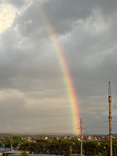 The image depicts a rainbow stretching across the sky above Zav'yalovo, Russia. The sky is overcast with grey clouds. Power lines and utility poles run across the upper right portion of the frame. Below the rainbow on the right, houses and trees are visible, forming a residential or suburban landscape. No people are visible in the image.