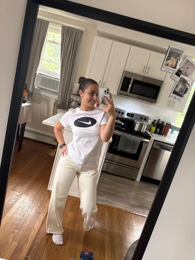 A young woman with her hair pulled up in a bun, wearing a white Nike t-shirt and cream-colored wide-leg pants, stands in a kitchen and takes a selfie in a large framed mirror. She is smiling and looking at her phone, which she holds in her right hand. Her left hand is on her hip. The kitchen features white cabinets, stainless steel appliances including a microwave and oven, and a tiled backsplash. There is a window with gray curtains and an air conditioning unit. On the refrigerator door, several instant photos are attached. The floor is made of polished wood.