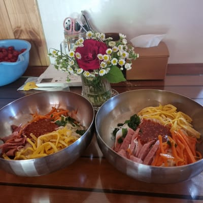 Two metal bowls filled with bibimbap are arranged on a wooden table. The bibimbap in both bowls appears to consist of noodles, shredded carrots, sliced ham, scrambled eggs, and a dollop of red sauce. A bouquet of small white flowers and a red rose in a glass vase sits behind the bowls, as does a wooden tissue box. To the left of the bowls, a blue plastic container filled with small red berries is visible. A calculator and a yellow decorative object are on the table in front of the tissue box. The background shows a white wall. The scene suggests a meal setting in a casual dining establishment or home in Dunnae-myeon, South Korea.