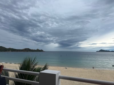 The image captures a view of the beach in Song Cau, Vietnam. The foreground features a white railing with a palm frond extending from the left side. Beyond the railing, a sandy beach stretches towards the ocean. The water is a calm, turquoise blue, with small waves lapping at the shore. A few distant boats are visible on the water. In the mid-ground, two people are standing near the water's edge. The background consists of a hilly coastline with vegetation, and the horizon is marked by a hazy sky filled with dramatic grey clouds. The overall impression is one of a serene, slightly overcast day at the beach.