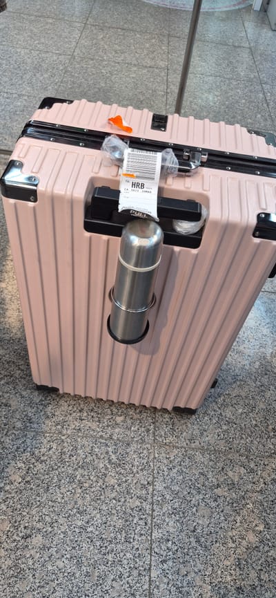 A light pink, ribbed suitcase is positioned upright on a speckled gray tiled floor. A metallic thermos is secured to the side of the suitcase with a ring holder. An airline baggage tag is attached to the suitcase handle, displaying text including "TO: HRB", "CA 1621 20MAR", and "VIA". The suitcase has black corner protectors and a black handle. The background shows more of the tiled floor and a metal pole. The scene is indoors, likely an airport terminal, given the baggage tag. There are no people visible in the image.